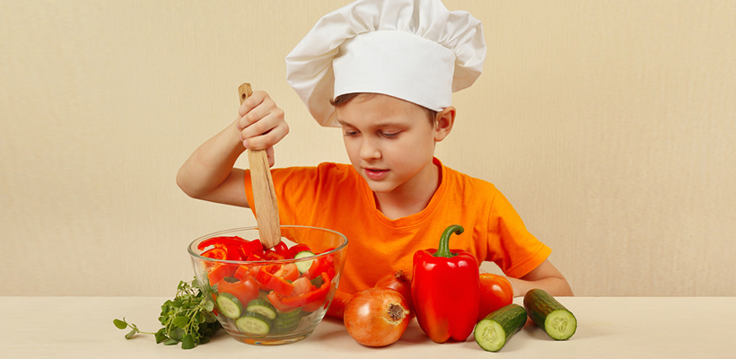 Little boy in chefs hat mixing the vegetables in a bowl with salad